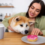Mulher segurando um cachorro enquanto ele observa um donut sobre um prato na mesa.