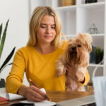 Mulher loira de suéter amarelo sentada à mesa de trabalho em casa, escrevendo em um caderno com lápis enquanto segura no colo um pequeno cachorro Yorkshire Terrier fofo, com livros, plantas e laptop ao fundo.